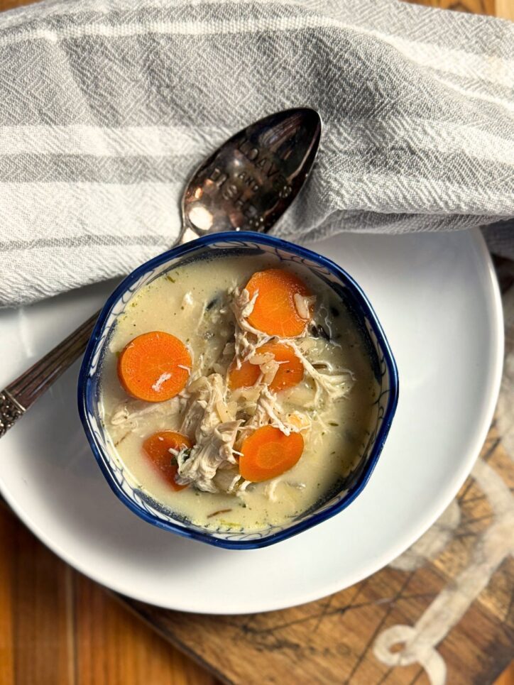 Bowl of creamy chicken soup with bright orange carrot slices and shredded chicken in a blue and white ceramic bowl, placed on a white plate with a vintage spoon and gray cloth napkin