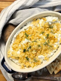 Dill pickle dip with crushed potato chips and fresh dill on top, in a casserole dish with a grey towel