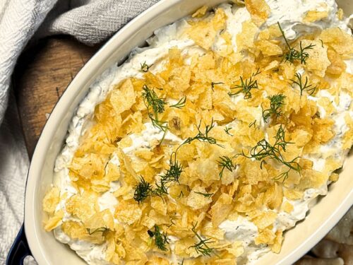 Dill pickle dip with crushed potato chips and fresh dill on top, in a casserole dish with a grey towel