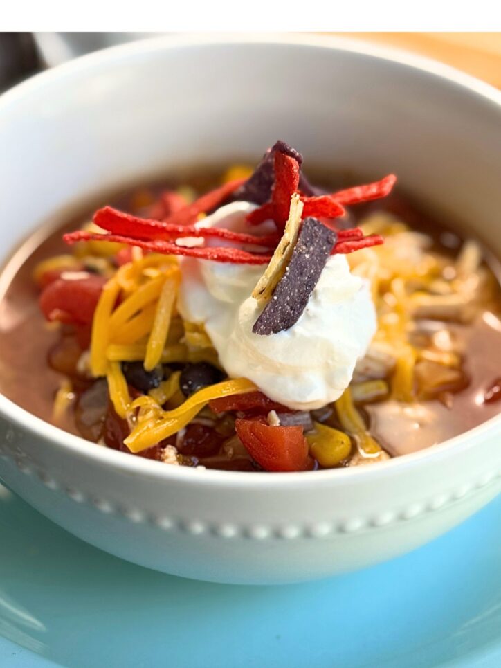 a close up view of the soup, topped with cheese, sour cream, and tortilla chips in a white bowl.