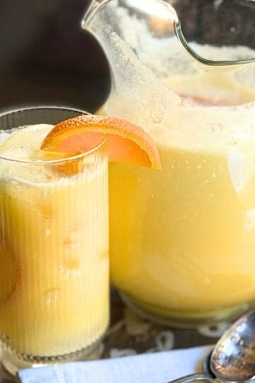 Glass pitcher and tumbler of Orange Coconut Refresher with an orange slice garnish, ice cubes, and a spoon on a wooden serving board.