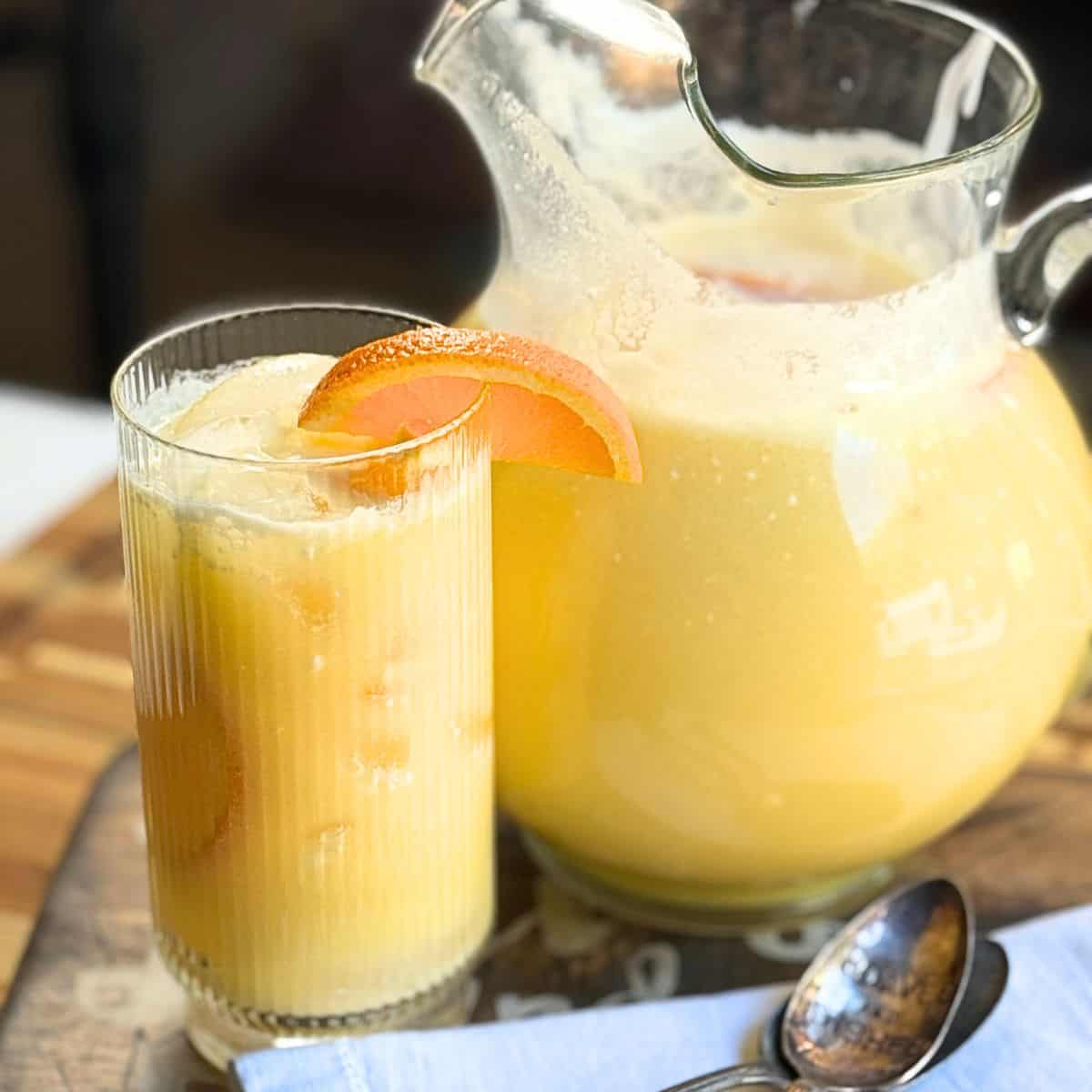 Glass pitcher and tumbler of Orange Coconut Refresher with an orange slice garnish, ice cubes, and a spoon on a wooden serving board.