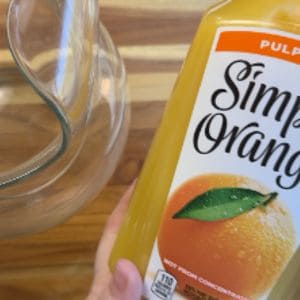 A glass pitcher on a wooden surface with a hand holding a bottle of Simply Orange juice with pulp.