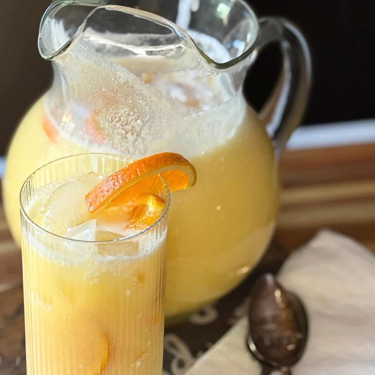 Glass pitcher of Orange Coconut Refresher with a serving glass, orange slice garnish, ice cubes, and a spoon on a wooden surface.