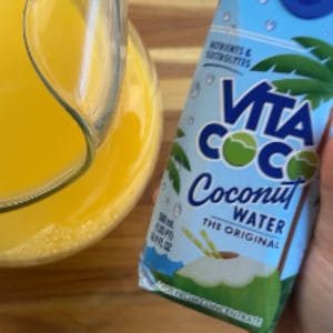 A glass pitcher filled with yellow-orange juice next to a hand holding a blue Vita Coco coconut water carton.