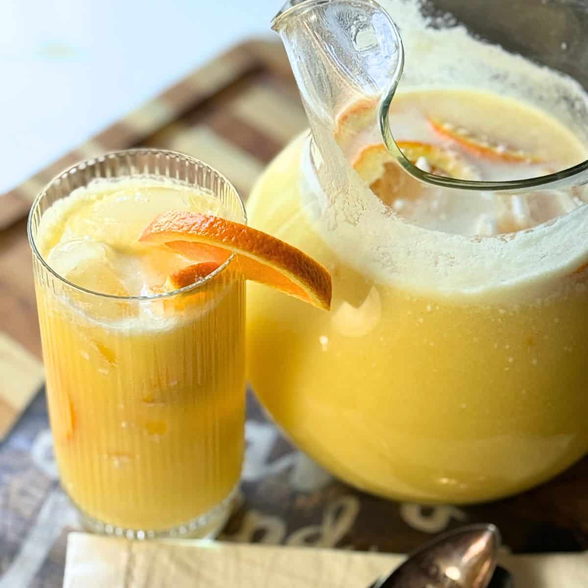 Close-up view of Orange Coconut Refresher in a pitcher and glass with orange slice garnish on a wooden tray.