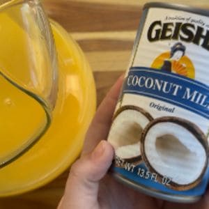 A glass pitcher containing orange-colored liquid next to a hand holding a can of Geisha coconut milk with a coconut illustration on the label.