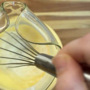 Close-up of a hand whisking yellow liquid in a glass pitcher, showing the whisk and the creamy mixture.